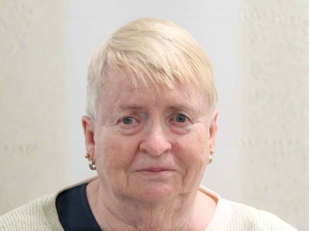 Close-up portrait of an older woman with short, light blonde hair and small gold earrings, wearing a cream sweater and neutral expression.