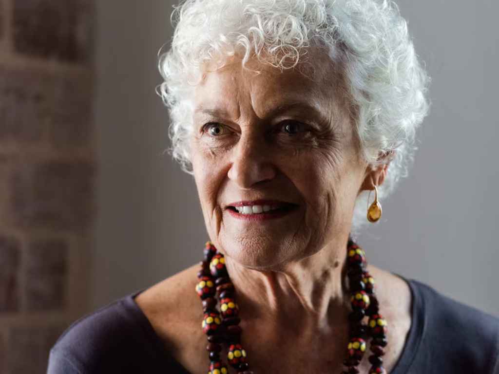 Portrait of an older woman with short white curly hair, smiling, wearing a colorful beaded necklace and amber earrings in a softly lit setting.