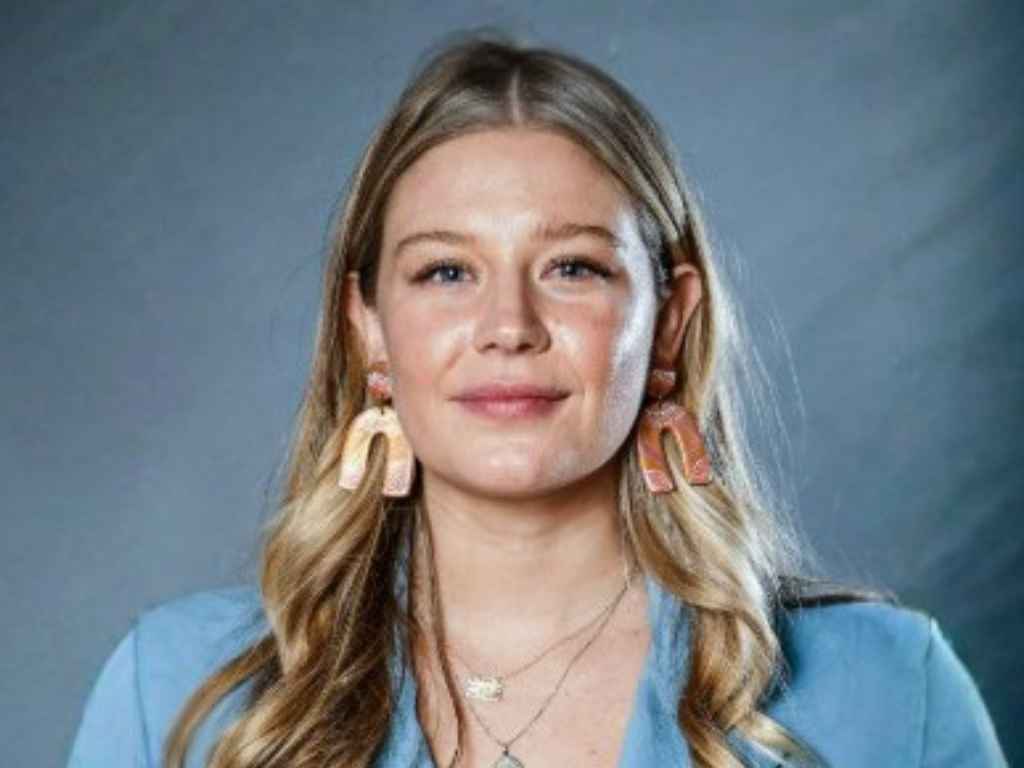 Portrait of a smiling woman with blonde hair wearing a blue blazer and pink arc earrings, facing the camera against a gray backdrop.