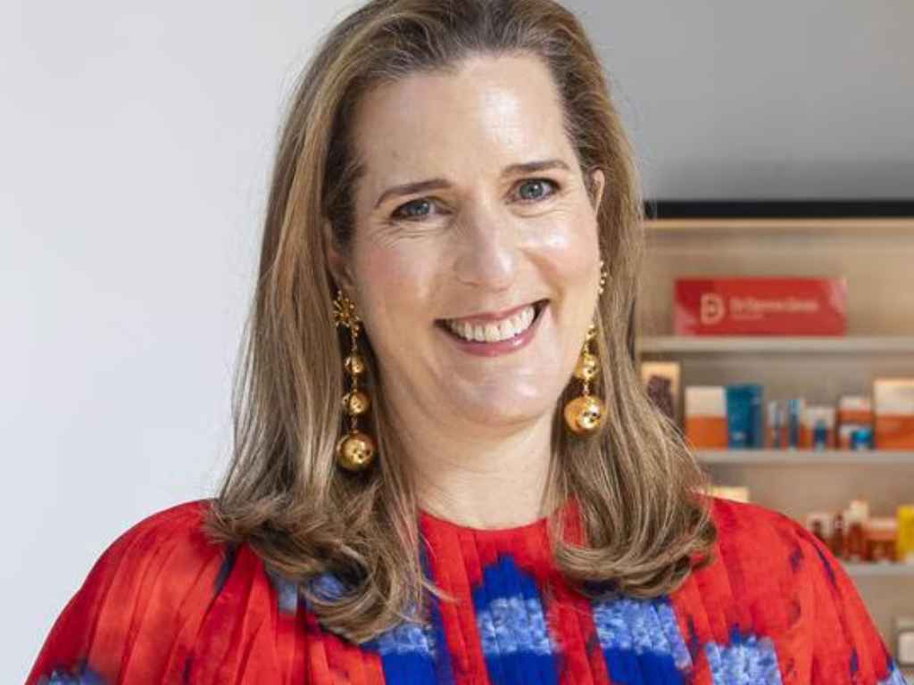 Smiling woman with shoulder-length blonde hair, wearing a red blouse and gold dangle earrings, in a bright room with shelves in the background.