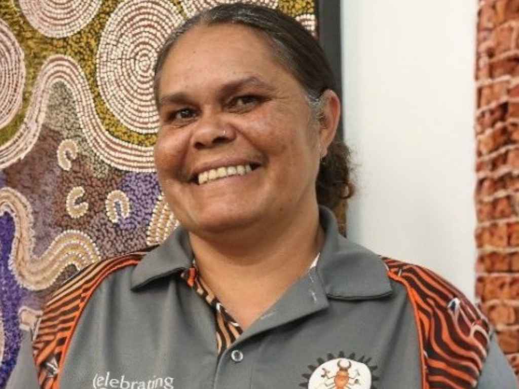 Smiling person wearing a gray polo with orange stripes, standing in front of Aboriginal art background and a badge on the chest.