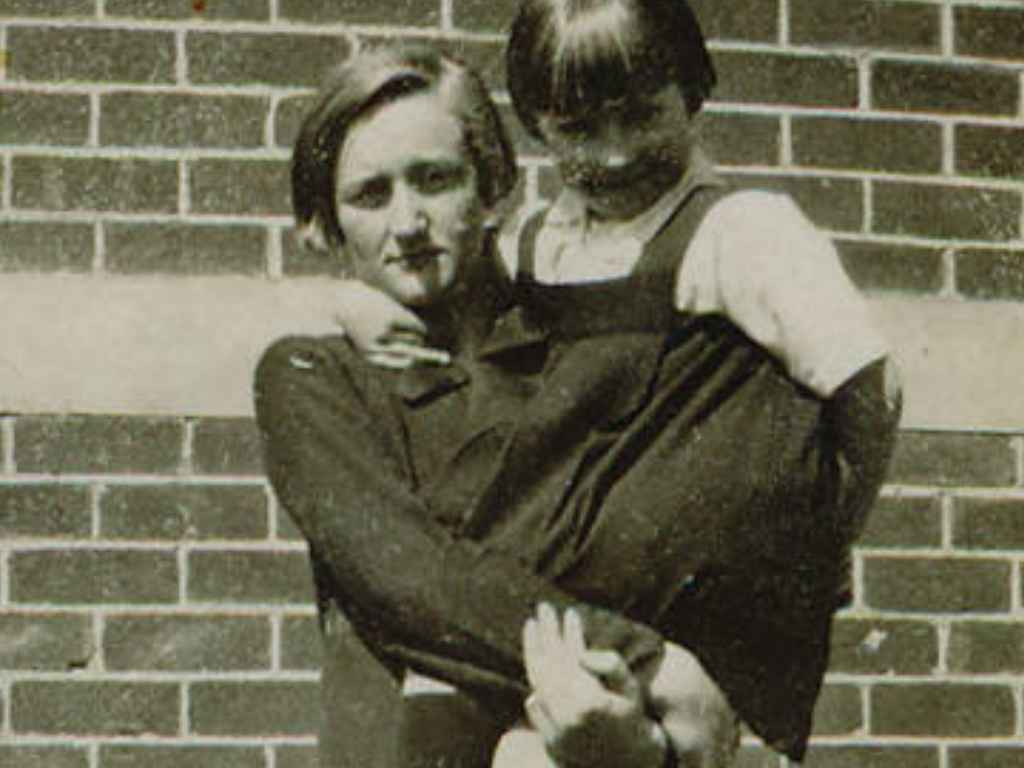 Woman carrying a child on her back, posing in front of a brick wall.