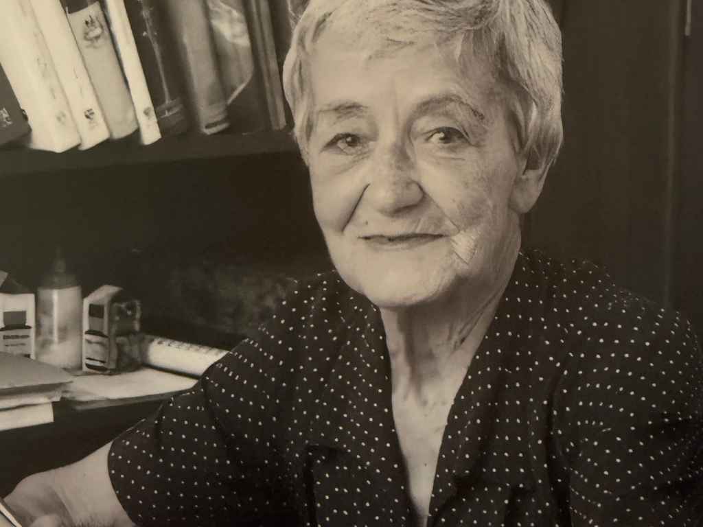 Portrait of an elderly woman with short gray hair wearing a black polka-dot blouse, seated at a desk with books and papers in the background.