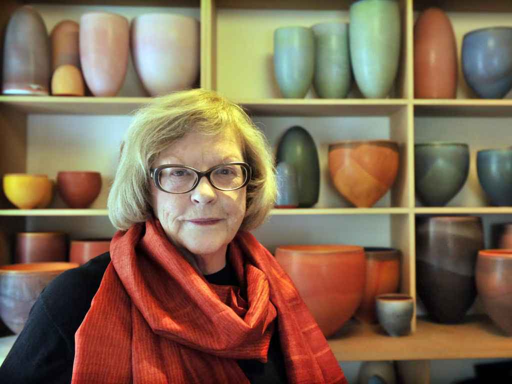 Smiling woman with glasses and a red scarf in front of shelves filled with colorful pottery.