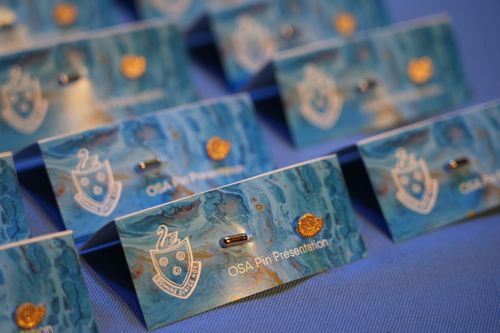 f_2_27548442 - St Hilda's Anglican School For Girls Row of blue presentation name badges with a crest and gold seal, labeled 'OSA Pin Presentation' on a blue tablecloth.