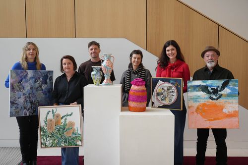 f_2_34208177 - St Hilda's Anglican School For Girls Six people stand in a gallery with their artworks: abstract painting, botanical illustration, ceramic sculpture on a pedestal, colorful woven vase, circular artwork, and a seascape with a bird.