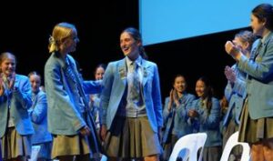 Group of students in blue blazers and gray skirts on a stage, clapping and chatting during a ceremony