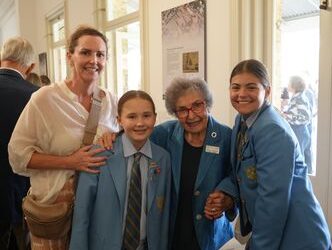 f_2_34489984 - St Hilda's Anglican School For Girls Two schoolgirls in blue blazers pose with an elderly woman and a smiling woman in a white top in a bright hallway.