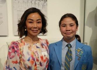 f_2_34490025 - St Hilda's Anglican School For Girls Two people posing indoors: a woman in a colorful floral dress beside a student in a blue blazer with a striped tie and badges.
