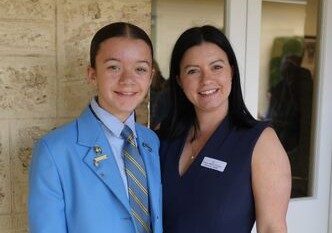 f_2_34490079 - St Hilda's Anglican School For Girls Smiling student in a light blue blazer and striped tie posing with a woman in a navy dress outside a brick building.
