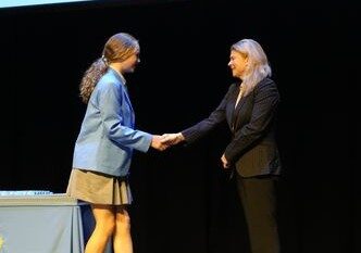 f_2_34490142 - St Hilda's Anglican School For Girls Two women shake hands on a stage, one in a blue blazer and beige skirt, the other in a black suit beside a table on the left.