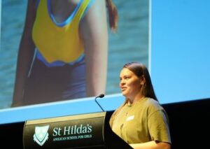 Woman in a yellow top and pink pants speaks at a black podium labeled 'St Hilda's Anglican School for Girls' on stage, with a large screen behind her showing a close-up image of someone wearing a yellow outfit.