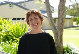 Smiling woman with short curly hair in a black top standing outdoors beside a railing, with trees and a yellow building in the background.