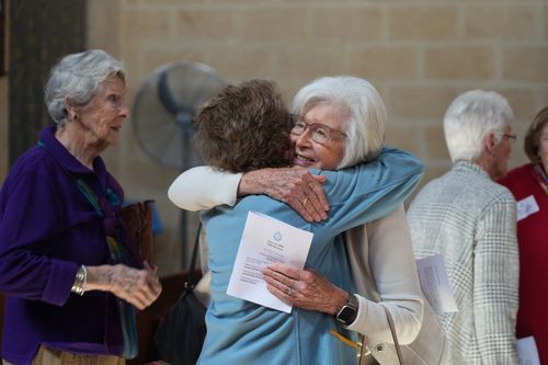f_2_34547679 - St Hilda's Anglican School For Girls Two elderly women share a warm hug at a social gathering, one holding a program flyer.