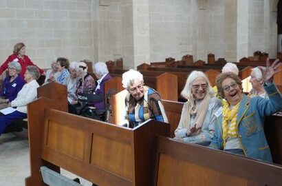 f_2_34547691 - St Hilda's Anglican School For Girls Group of elderly women seated in church pews inside a stone church; several smile at the camera, one waves.