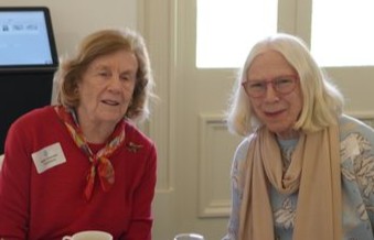 f_2_34547725 - St Hilda's Anglican School For Girls Two elderly women sit at a table in a bright room, smiling. Left woman wears a red sweater with a name tag and scarf; right woman wears glasses and a light scarf.