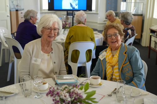f_2_34547729 - St Hilda's Anglican School For Girls Two smiling elderly women sit at a round table with tea cups and a flower centerpiece during a social gathering.