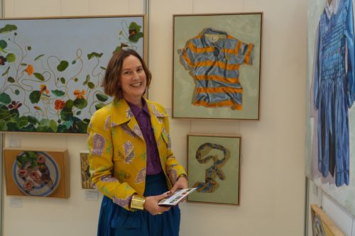 f_2_34611331 - St Hilda's Anglican School For Girls Smiling woman in a yellow patterned jacket standing in an art gallery, holding pamphlets, with framed artworks on the wall around her.