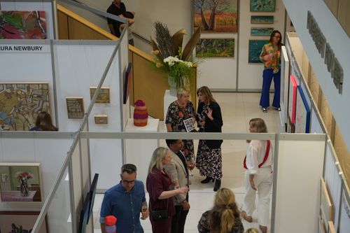 f_2_34611340 - St Hilda's Anglican School For Girls Group of visitors chat and stroll through an art fair with white partition booths and colorful paintings on the walls at a gallery event.