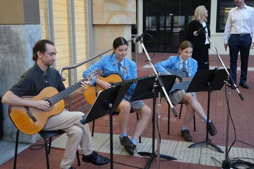 f_2_34611344 - St Hilda's Anglican School For Girls Three musicians playing guitars outdoors: a man and two girls in blue jackets sit on chairs with music stands and microphones on a brick plaza.