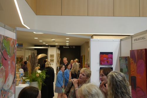 f_2_34611345 - St Hilda's Anglican School For Girls Crowd of visitors at an indoor art exhibition, white display booths and colorful paintings on the walls as attendees mingle.
