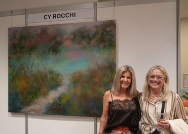 f_2_34614918 - St Hilda's Anglican School For Girls Two smiling women pose in front of a large abstract landscape painting at a booth labeled CY ROCCHI in an art fair.