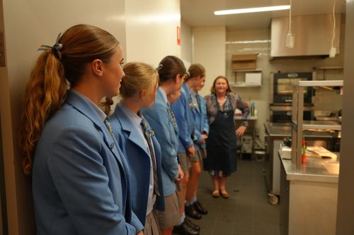 f_2_34614919 - St Hilda's Anglican School For Girls Six students in matching blue jackets stand in a commercial kitchen, listening to a woman in an apron giving instructions.
