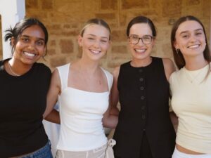 Four smiling young women standing side by side indoors, arms around each other.