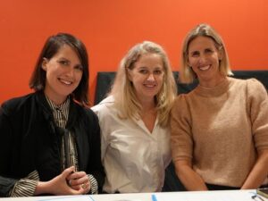 Three women posing together in an office, smiling at the camera with an orange wall behind them.
