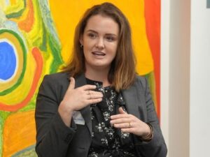 Woman in a dark gray blazer speaks, hands gesturing, in front of a colorful abstract painting in a gallery or office setting.