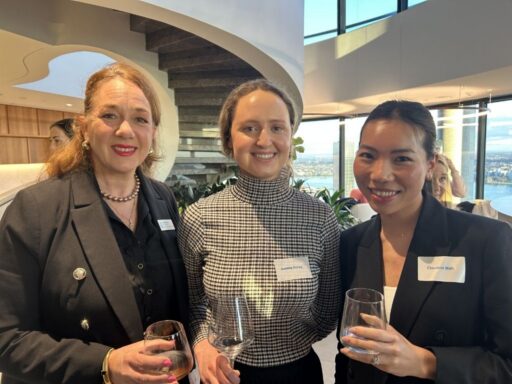 Three women in business attire pose for a photo at a high-rise networking event, each holding a wine glass in a modern lounge setting with a city view behind them.