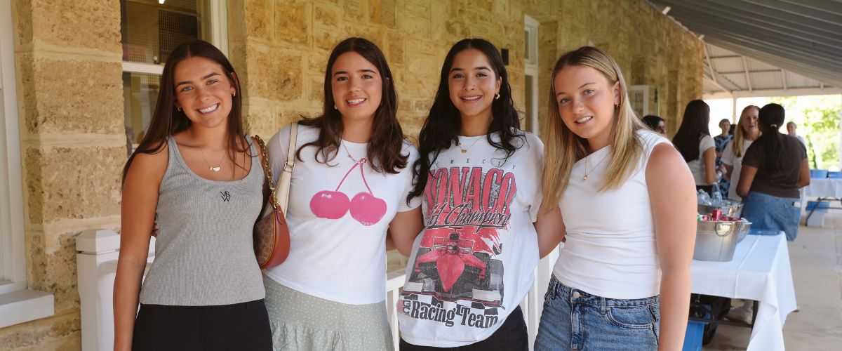 Four young women standing together, smiling, outside a stone building at an event.