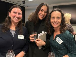 Three women at a networking event smiling for the camera; the center woman holds a glass and a notebook beside the others with name tags visible.