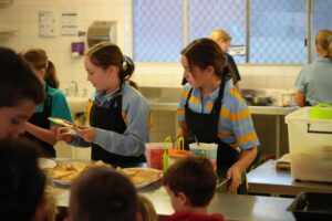 STH00475 1 - St Hilda's Anglican School For Girls Children in a school kitchen wearing aprons serve pastries at a counter; pie trays sit nearby as others chat in the background.