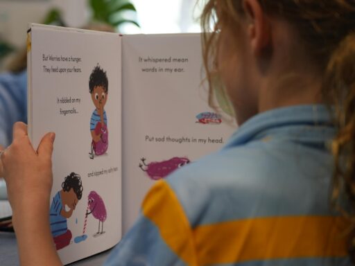 Child sits at a table in a classroom, reading a colorful illustrated book with pages open to cartoons.