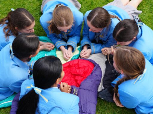 Overhead view of eight girls in matching blue uniforms sitting in a circle on the grass, hands joined over a colorful sleeping bag in the center.
