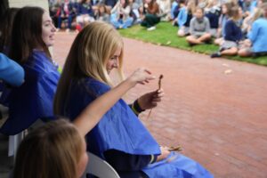 STH03786 1 1 - St Hilda's Anglican School For Girls Two women in blue ponchos sitting outdoors, smiling while handling a small stick-like object as a crowd watches nearby on a brick path and grass.