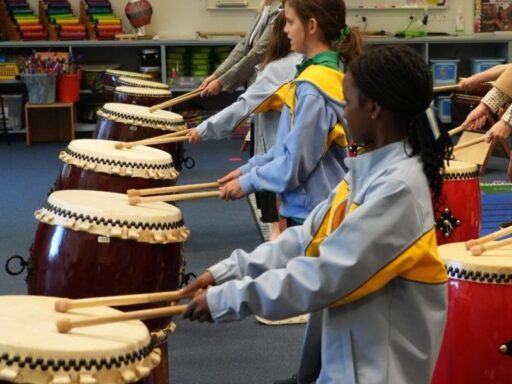 Children and a teacher playing taiko drums in a classroom, each student striking a drum with wooden sticks.